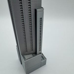 May include: A gray, 3D-printed model of a skyscraper. The building has many windows and a rectangular shape. The model is detailed, showing the structure's facade and architectural design. The building has a smaller tower attached to the main structure.