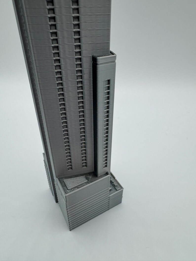 May include: A gray, 3D-printed model of a skyscraper. The building has many windows and a rectangular shape. The model is detailed, showing the structure's facade and architectural design. The building has a smaller tower attached to the main structure.