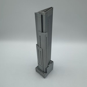 May include: A gray, 3D-printed model of a skyscraper. The building has a rectangular base, a tall, slender main structure with many windows, and a flat roof. The model is a solid, monochromatic gray.