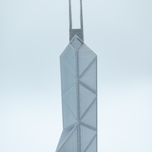 May include: A gray, 3D-printed architectural model of a skyscraper. The building features a geometric design with triangular and rectangular shapes. The model has a tall, slender profile with two vertical extensions at the top.