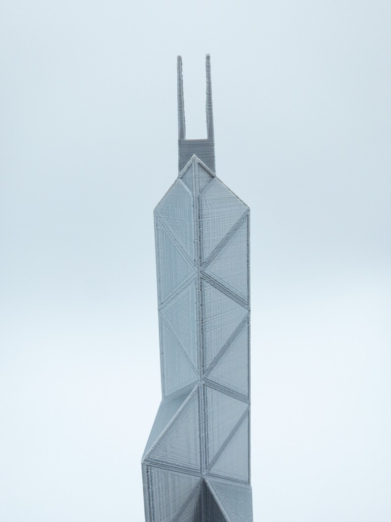 May include: A gray, 3D-printed architectural model of a skyscraper. The building features a geometric design with triangular and rectangular shapes. The model has a tall, slender profile with two vertical extensions at the top.