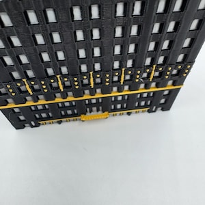 Op de afbeelding: Een zwart en geel architectonisch model met een rasterachtige structuur, met rechthoekige openingen en horizontale gele accenten. Het model lijkt een miniatuurweergave van een gebouw of structuur te zijn.