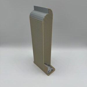 May include: A tan and gray miniature skyscraper model. The building has a rectangular shape with a grid-like texture on the sides. The top of the building is gray, and the base is tan. The model is standing upright on a white surface.