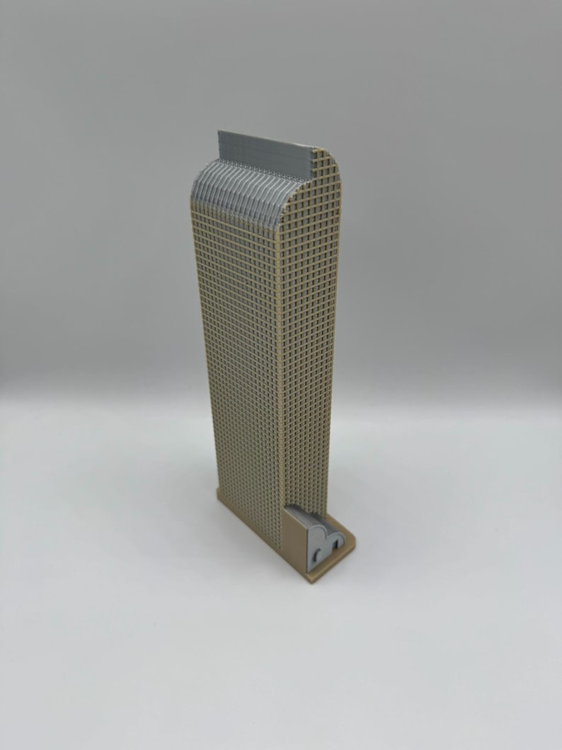 May include: A tan and gray miniature skyscraper model. The building has a rectangular shape with a grid-like texture on the sides. The top of the building is gray, and the base is tan. The model is standing upright on a white surface.