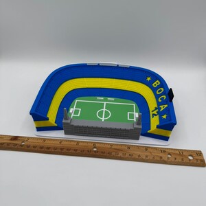 May include: A miniature stadium model with blue and yellow seating, a green field, and a grey structure. The model has the text "BOCA 12" on the side. A wooden ruler is placed next to the model.