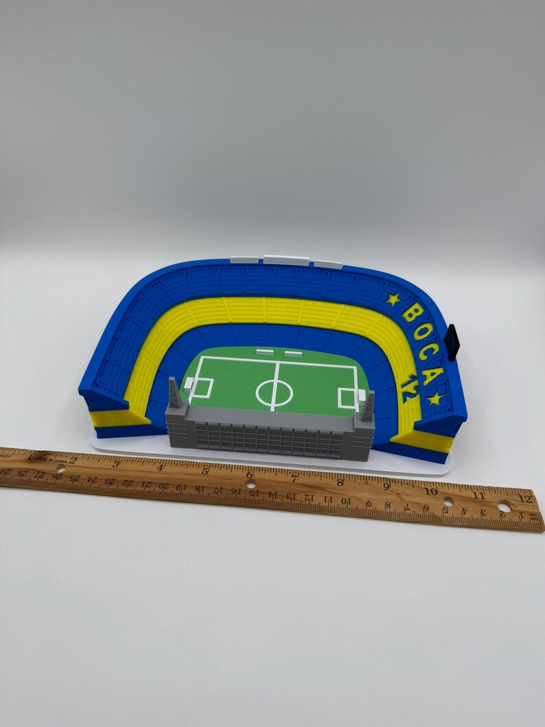 May include: A miniature stadium model with blue and yellow seating, a green field, and a grey structure. The model has the text "BOCA 12" on the side. A wooden ruler is placed next to the model.