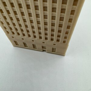 Pu&ograve; includere: Un modello di edificio alto di colore beige stampato in 3D con un motivo a griglia sulla facciata e una fila di finestre alla base.