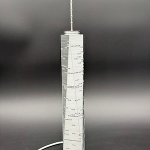 May include: A gray model of a tall skyscraper with a spire. The building has many windows and a detailed facade. The model is made of a light gray material and stands on a flat base. A white cable is connected to the base.