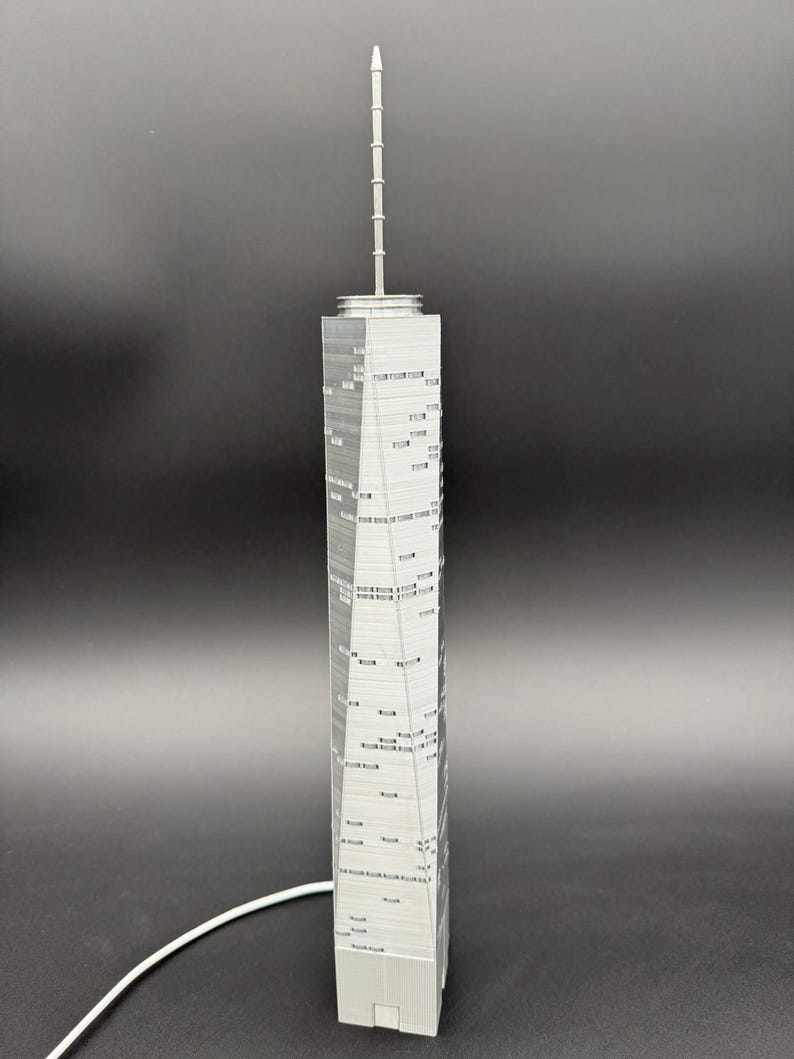 May include: A gray model of a tall skyscraper with a spire. The building has many windows and a detailed facade. The model is made of a light gray material and stands on a flat base. A white cable is connected to the base.