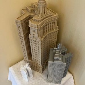 Op de afbeelding: Drie miniatuurgebouwenmodellen, een beige met een koepel, een grijs met een koepel en een wit met een koepel, op een wit tafelkleed.