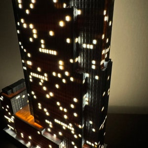 May include: A detailed model of a tall skyscraper with many windows, lit up with small white lights. The model is made of a dark gray material and has a brown base.