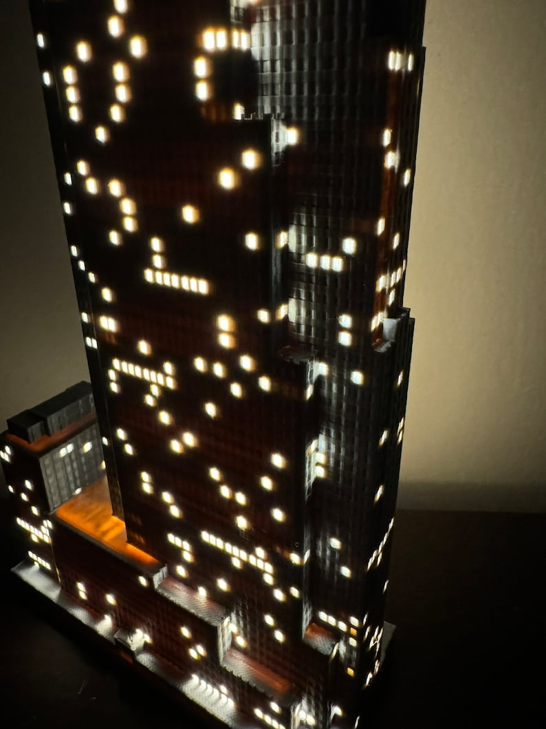 May include: A detailed model of a tall skyscraper with many windows, lit up with small white lights. The model is made of a dark gray material and has a brown base.