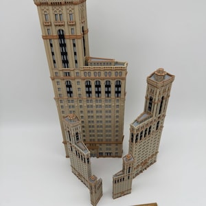 May include: Four beige miniature building models with architectural details, including windows and decorative elements. A wooden ruler is placed near the buildings. The buildings vary in size, with the tallest one being the largest.