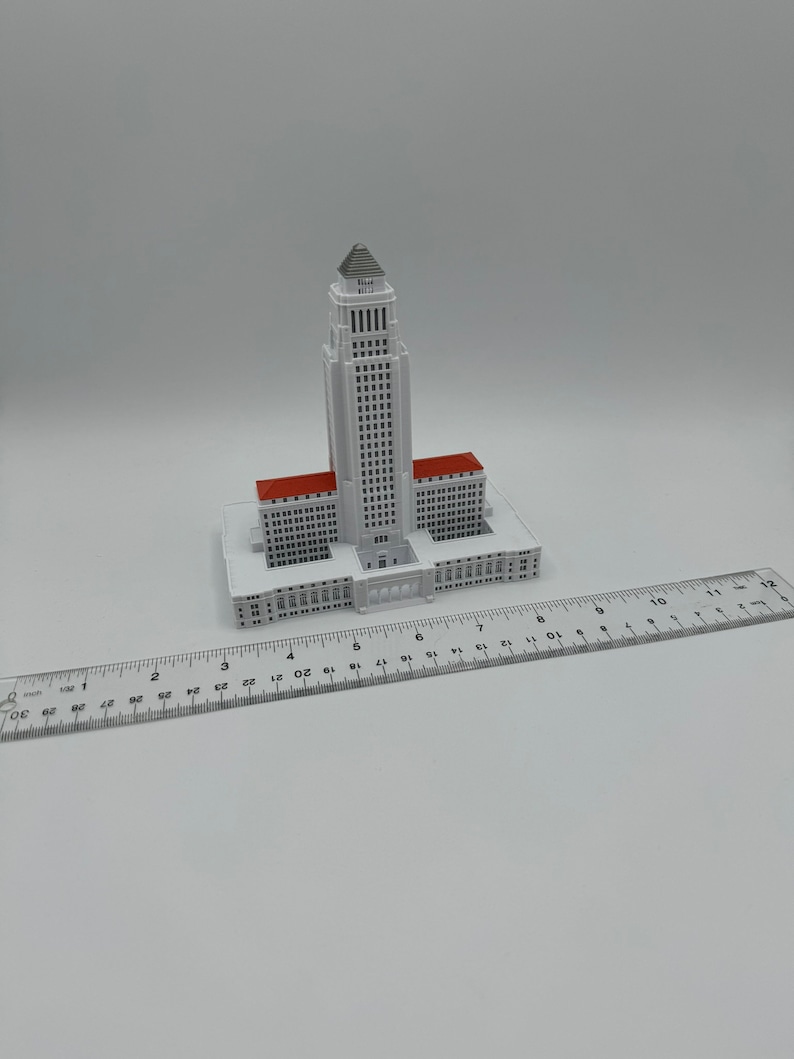 May include: A white miniature model of the Los Angeles City Hall building with a red roof. The model is approximately 6 inches tall and is sitting on a white surface next to a ruler.