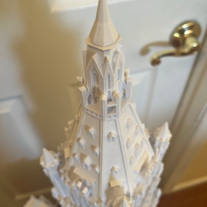 May include: A white 3D printed model of a tall, ornate building with a pointed spire. The building has many windows and intricate details.