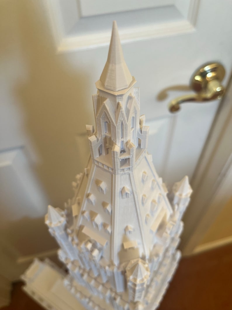 May include: A white 3D printed model of a tall, ornate building with a pointed spire. The building has many windows and intricate details.