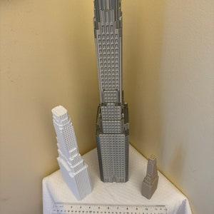 May include: Three 3D printed model buildings, one tall and gray, one medium and white, and one small and tan. The buildings are on a white tablecloth with a ruler in the foreground.