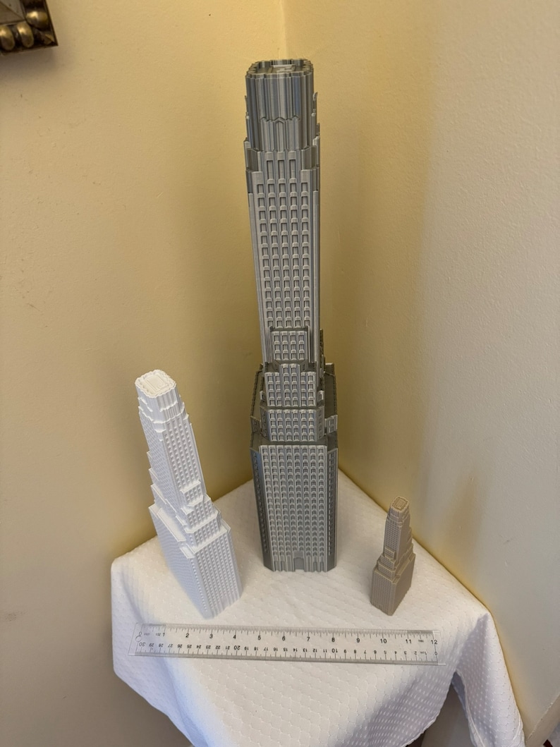 May include: Three 3D printed model buildings, one tall and gray, one medium and white, and one small and tan. The buildings are on a white tablecloth with a ruler in the foreground.