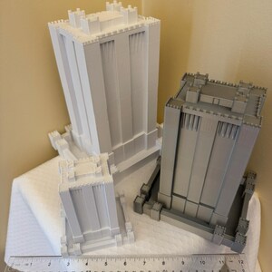 May include: Two 3D printed models of buildings with a castle-like design. The buildings are white and gray and are sitting on a white surface. A ruler is in the foreground.
