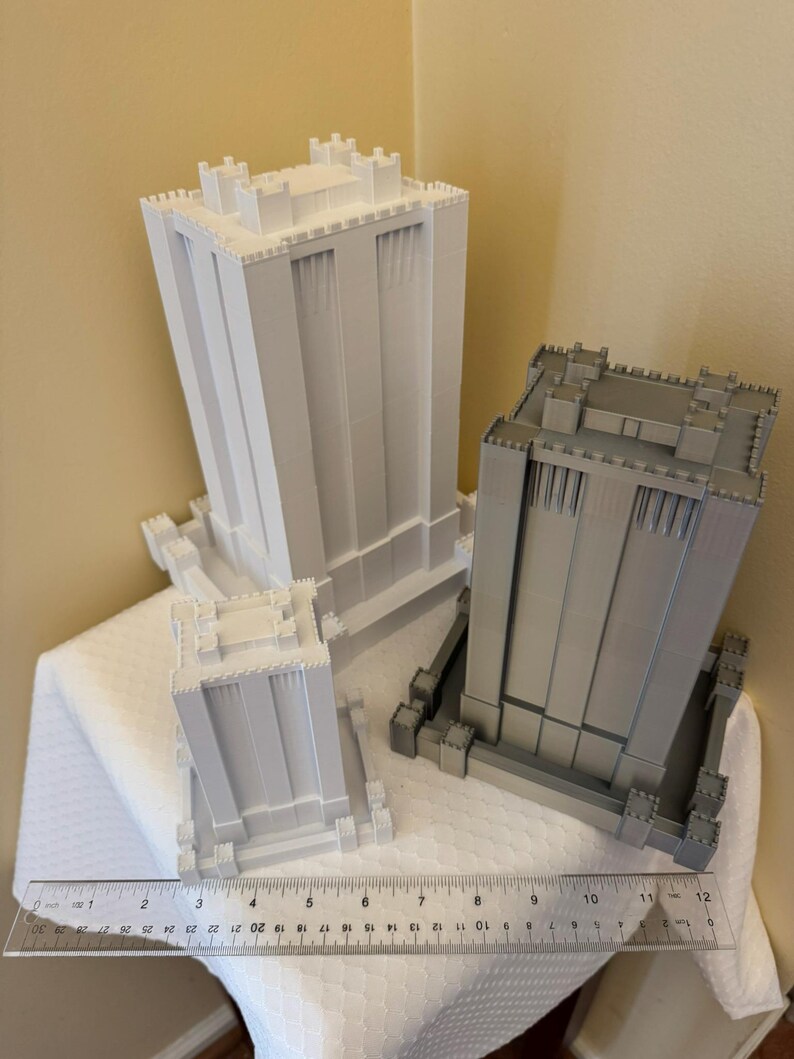 May include: Two 3D printed models of buildings with a castle-like design. The buildings are white and gray and are sitting on a white surface. A ruler is in the foreground.