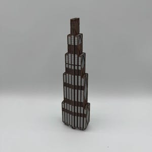 May include: A vertical architectural model of a skyscraper featuring a stepped, tiered design. The structure is composed of dark brown framing with light grey rectangular panels on each level, creating a repeating geometric pattern throughout.
