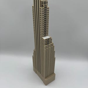 May include: A beige-colored miniature skyscraper model with a detailed facade. The building has a tall, slender main structure and a shorter, wider base. The model is a representation of a city building.