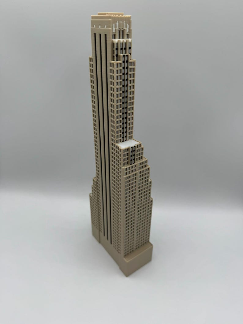 May include: A beige-colored miniature skyscraper model with a detailed facade. The building has a tall, slender main structure and a shorter, wider base. The model is a representation of a city building.
