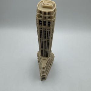 Op de afbeelding: Een beige miniatuurmodel van een hoge wolkenkrabber met een plat dak en een decoratieve bovenkant. Het model is gemaakt van plastic en heeft een gedetailleerde gevel.