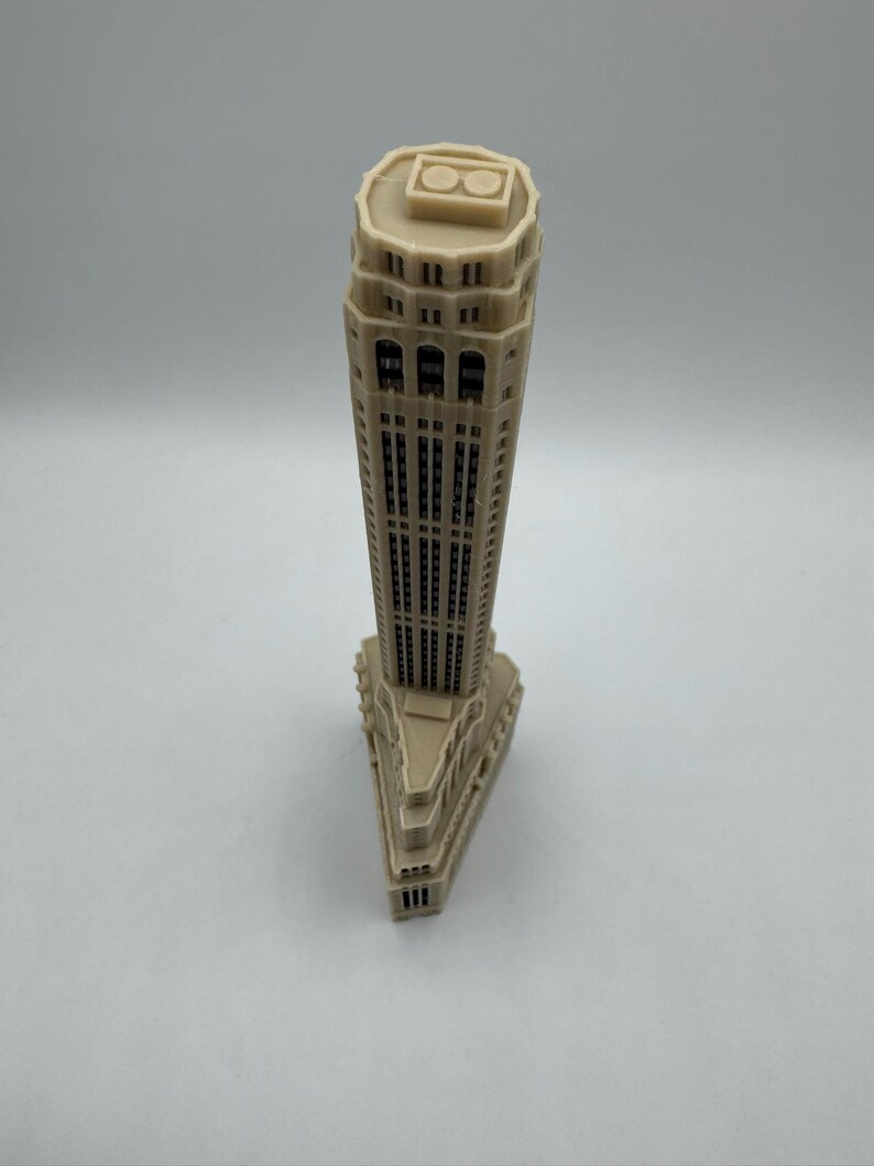 Op de afbeelding: Een beige miniatuurmodel van een hoge wolkenkrabber met een plat dak en een decoratieve bovenkant. Het model is gemaakt van plastic en heeft een gedetailleerde gevel.