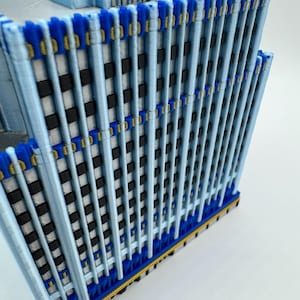May include: A 3D-printed model of a skyscraper. The building features a light blue facade with vertical supports, accented by dark blue and gold details. The windows are represented by black and white squares, creating a realistic architectural design.