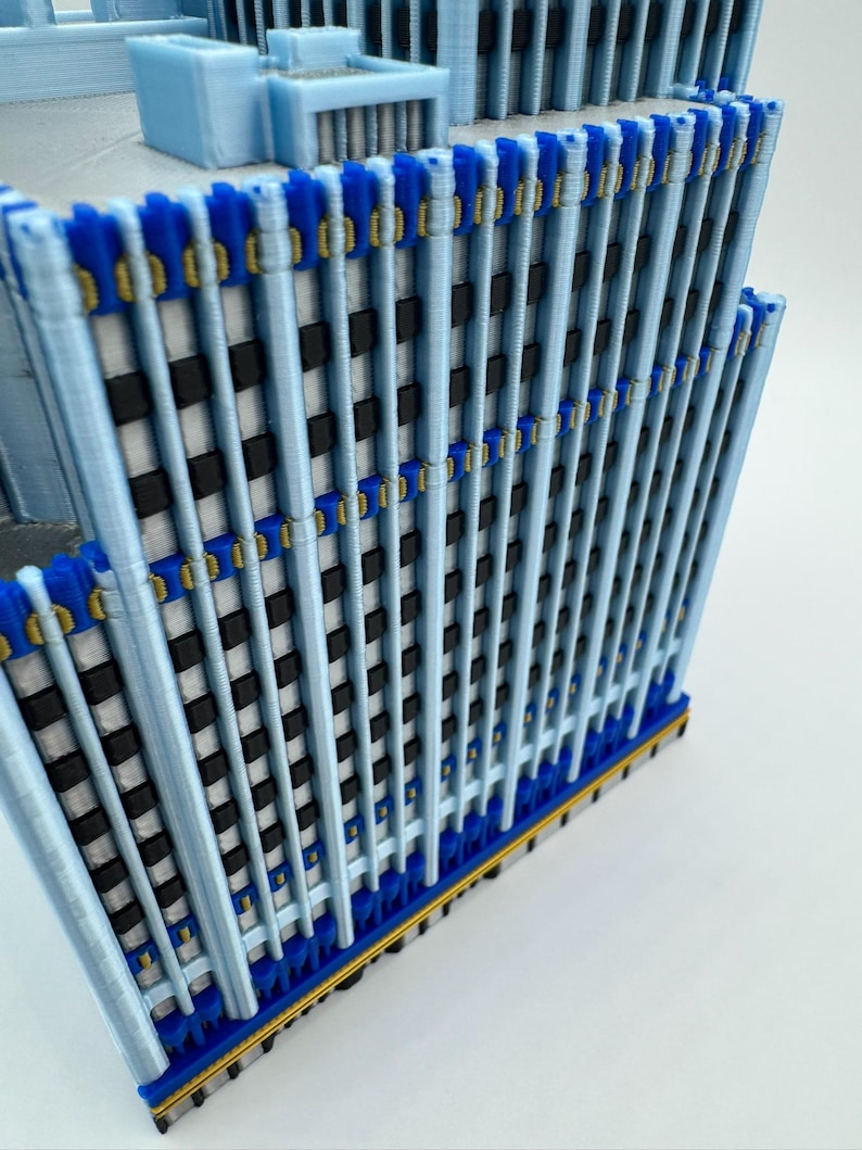 May include: A 3D-printed model of a skyscraper. The building features a light blue facade with vertical supports, accented by dark blue and gold details. The windows are represented by black and white squares, creating a realistic architectural design.