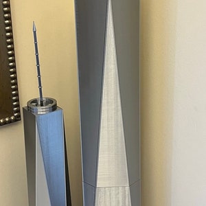 May include: Two silver-colored 3D printed models of skyscrapers with pointed tops. The larger model is taller than the smaller model. Both models are sitting on a wooden table.