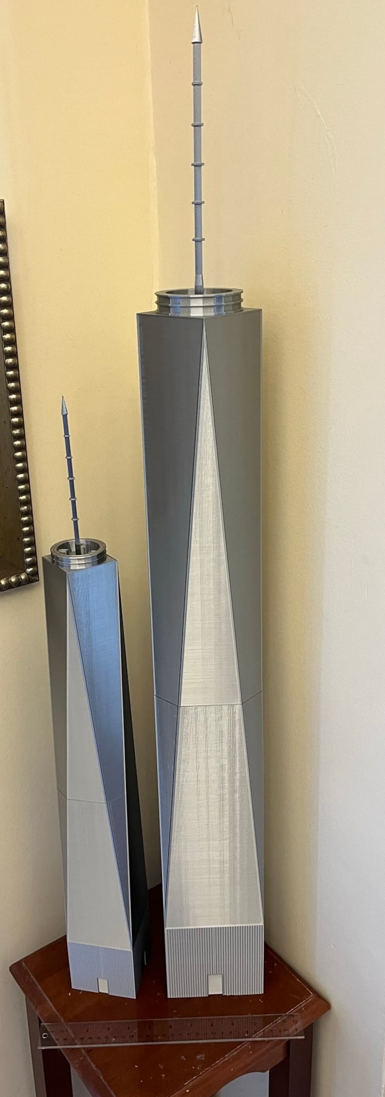 May include: Two silver-colored 3D printed models of skyscrapers with pointed tops. The larger model is taller than the smaller model. Both models are sitting on a wooden table.