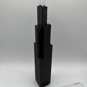 May include: A black 3D printed model of a skyscraper with a white antenna on top. The model is about 7 inches tall and is sitting on a white surface.
