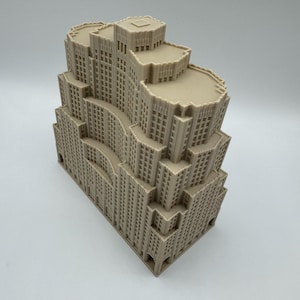 May include: A beige architectural model of a multi-tiered skyscraper. The model has detailed windows and a stepped design, with a flat roof. The building's facade has a textured appearance, and the overall design is reminiscent of Art Deco architecture.