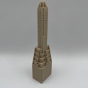 May include: A tan-colored architectural model of a skyscraper. The building has a detailed facade with many windows and a tiered design. The top of the building has intricate detailing. The model is set against a plain, light background.