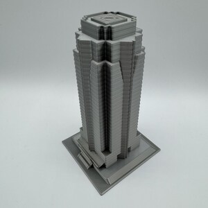 May include: A gray, 3D-printed model of a skyscraper. The building has a rectangular base and a multi-tiered design, with vertical lines creating a textured facade. The top of the building features a square, recessed design.