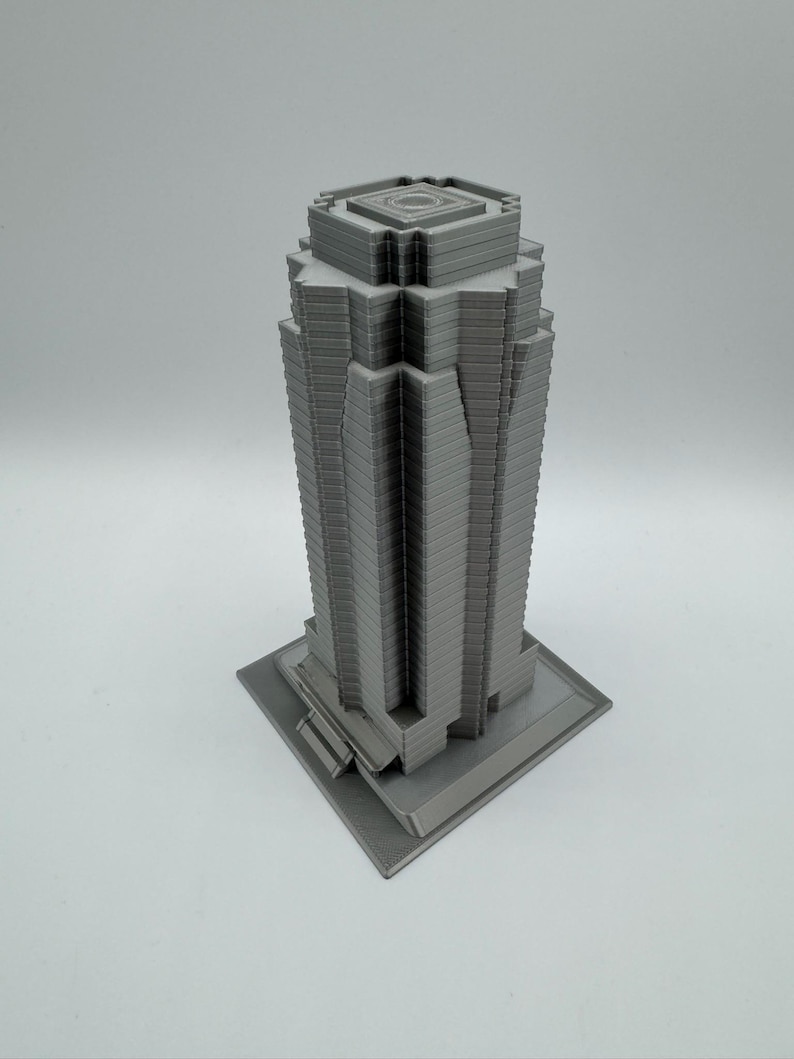 May include: A gray, 3D-printed model of a skyscraper. The building has a rectangular base and a multi-tiered design, with vertical lines creating a textured facade. The top of the building features a square, recessed design.