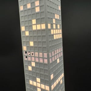 May include: A tall, white, 3D-printed model of a skyscraper with illuminated windows. The windows are lit with a warm, yellow light, creating a contrast against the white exterior. The building has a grid-like pattern.