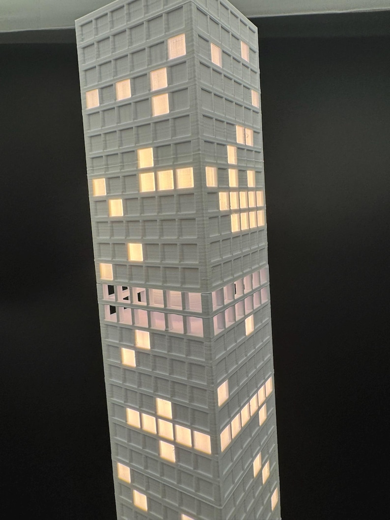 May include: A tall, white, 3D-printed model of a skyscraper with illuminated windows. The windows are lit with a warm, yellow light, creating a contrast against the white exterior. The building has a grid-like pattern.