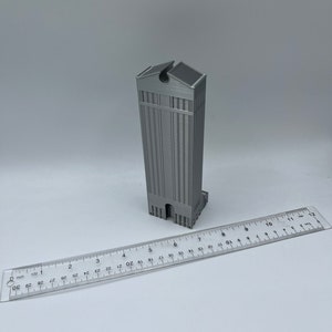 May include: A 3D printed model of a tall, gray building with a flat roof and a small opening at the top. The model is about 6 inches tall and is sitting on a white surface.