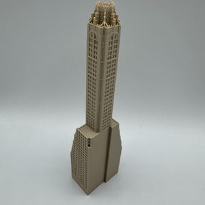May include: A beige architectural model of a skyscraper. The building features a detailed facade with numerous windows and a decorative top. The base of the building has a unique, angled design. The model is set against a plain white background.