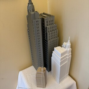 Pu&ograve; includere: Tre edifici modello stampati in 3D, uno grigio, uno bianco e uno beige, sono disposti su una tovaglia bianca. Un righello si trova in primo piano.
