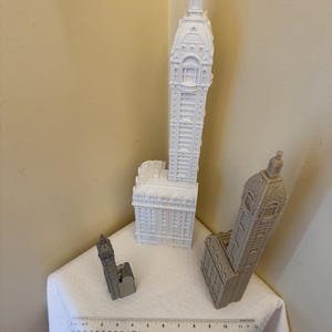 May include: Three white and gray miniature building models. The tallest building is white and has a pointed roof. The middle building is gray and has a flat roof. The smallest building is white and has a pointed roof. A ruler is in the foreground.
