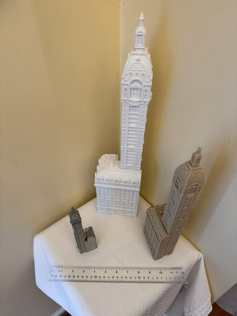 May include: Three white and gray miniature building models. The tallest building is white and has a pointed roof. The middle building is gray and has a flat roof. The smallest building is white and has a pointed roof. A ruler is in the foreground.