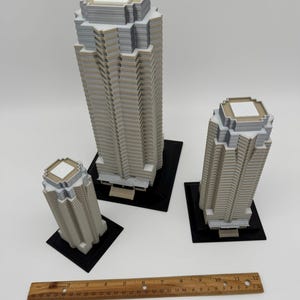 May include: Three miniature skyscraper models in varying sizes. The buildings are primarily light gray with white and dark gray accents. Each sits on a black square base. A wooden ruler is placed near the buildings.
