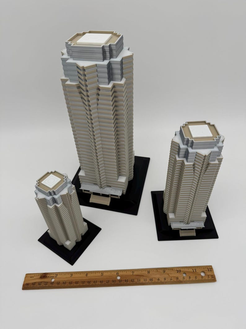 May include: Three miniature skyscraper models in varying sizes. The buildings are primarily light gray with white and dark gray accents. Each sits on a black square base. A wooden ruler is placed near the buildings.