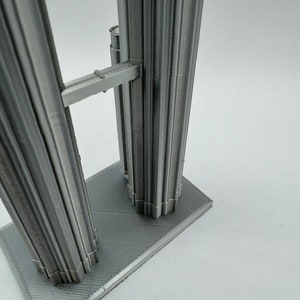 May include: A 3D printed model of a tall, gray building with a connecting bridge between two towers. The model is on a gray base.
