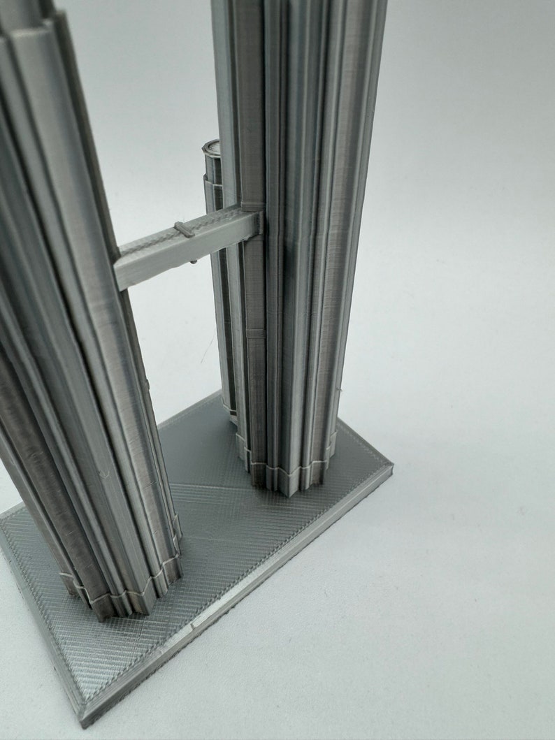 May include: A 3D printed model of a tall, gray building with a connecting bridge between two towers. The model is on a gray base.