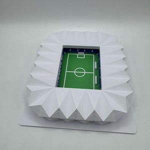 May include: A miniature white paper stadium model with a green field and blue accents. The stadium has a detailed design, including seating areas and a playing field. The text "[UNREADABLE]" is visible on the side of the stadium.
